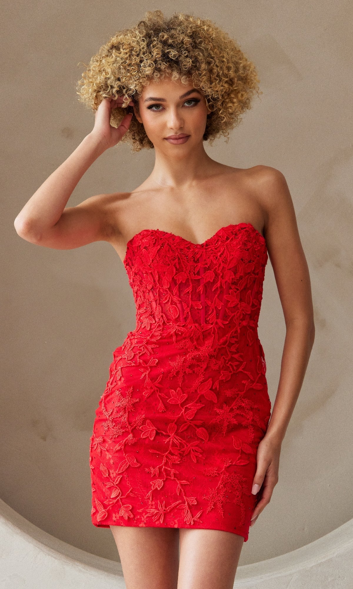 A woman wears the Embroidered-Lace Strapless Short Hoco Dress 7053S, a form-fitting red homecoming dress with a sweetheart neckline and floral embroidered lace, standing against a light background.